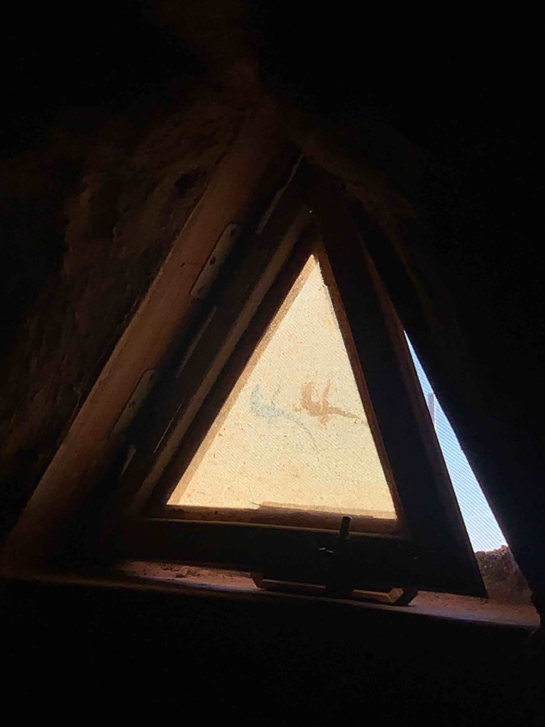 A small lizard and its shadow can be seen against the glass of a propped open triangular window.