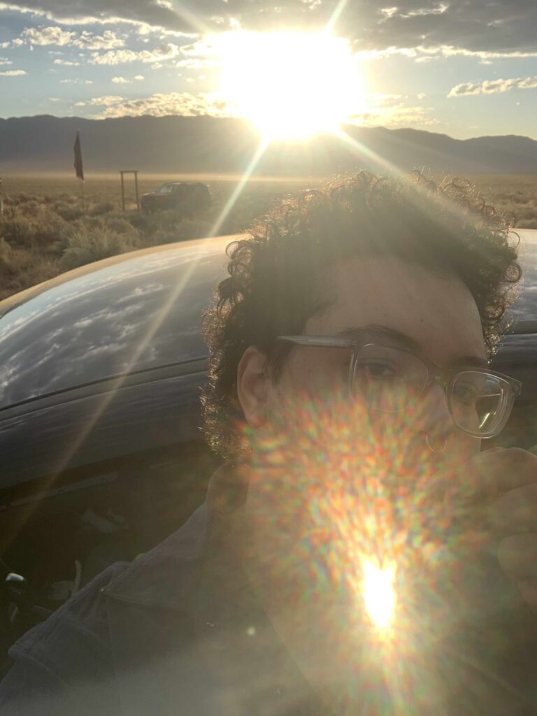 Azul brushes their teeth with a sunrise behind them. The sun's glare reflects in the camera, covering their face.