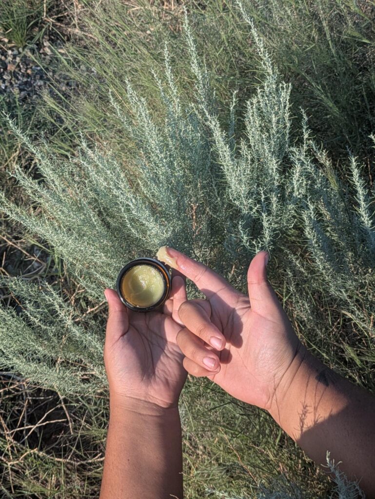 Azul's hands holding a jar of salve with a dollop on their finger. Sagebrush is in the background.