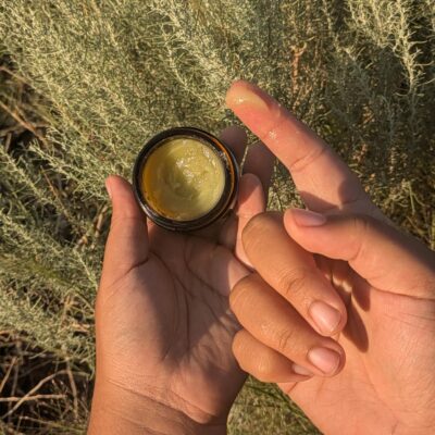 Azul's hands holding a jar of salve with a dollop on their finger. Sagebrush is in the background.